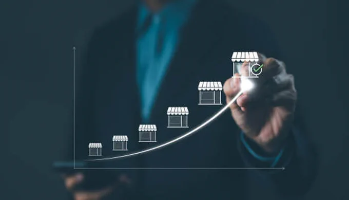 Image of a person in a suit using a smartphone, drawing a graph with small storefront icons rising along an upward curve, symbolizing franchise growth and success.