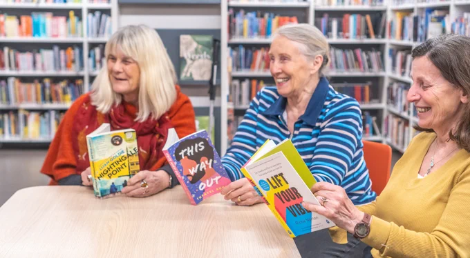 Photo of three older women sat at a table in a library holding books and smiling.