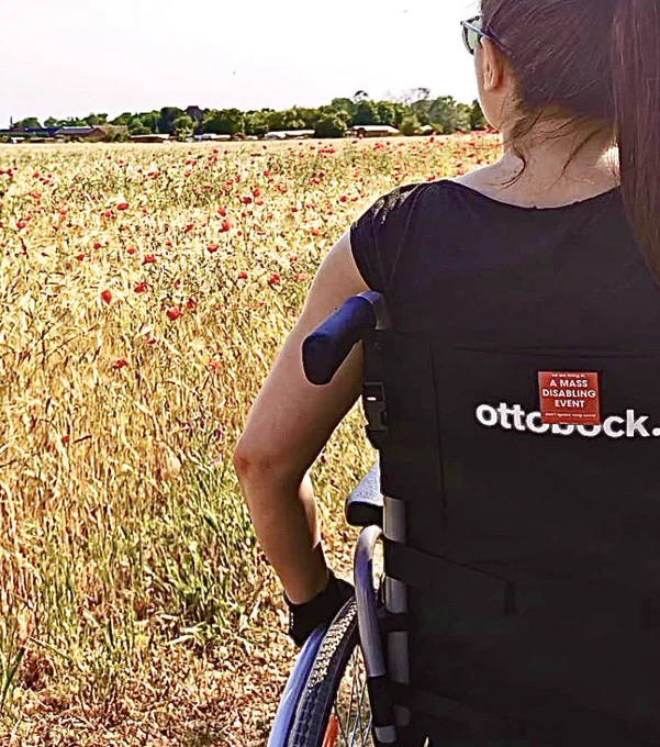 A picture of me sitting on a wheelchair. On the back of the wheelchair there’s an awareness sticker by @BerlinBuyers: “We’re living in a mass disabling event”