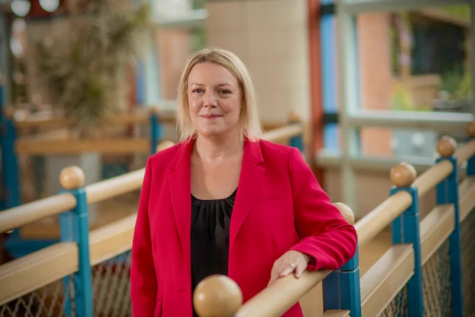 Professor Charlotte Summers with short blond hair is wearing a bright red blazer over a black top, standing in a modern, well-lit interior with blue and beige railings. The background features large windows and blurred foliage, creating an open and airy atmosphere.