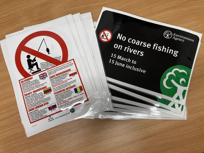 Stack of informational leaflets and signs on a wooden surface. The left stack features a "No Fishing" symbol with details in English and other languages. The right stack displays a "No coarse fishing on rivers – 15 March to 15 June inclusive" sign with the Environment Agency logo.