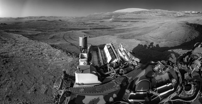 A Mars rover in the foreground, with a rocky desert landscape seen in panoramic format, a rover tracks