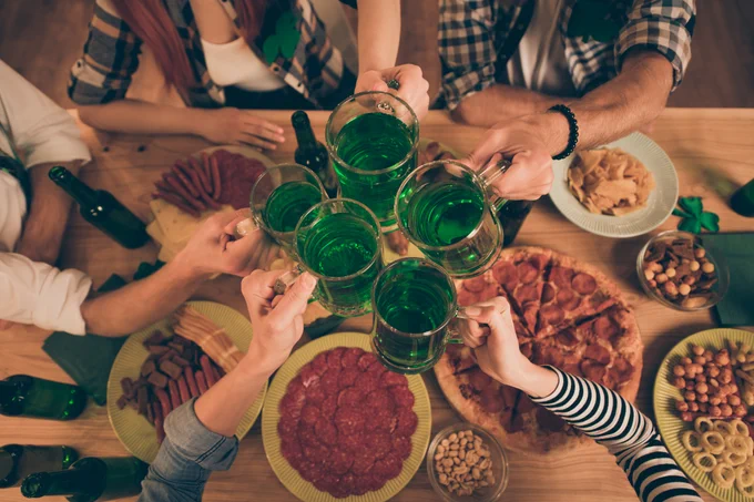 A group of people hold mugs of green beer over a table filled with various snacks, including pizza, pretzels, and sliced meats. The setting suggests a festive atmosphere, possibly celebrating St. Patrick's Day.