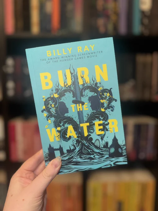 A hand holds a blue book titled "Burn the Water" by Billy Ray. The cover features ornate black designs. Blurred bookshelf in the background.