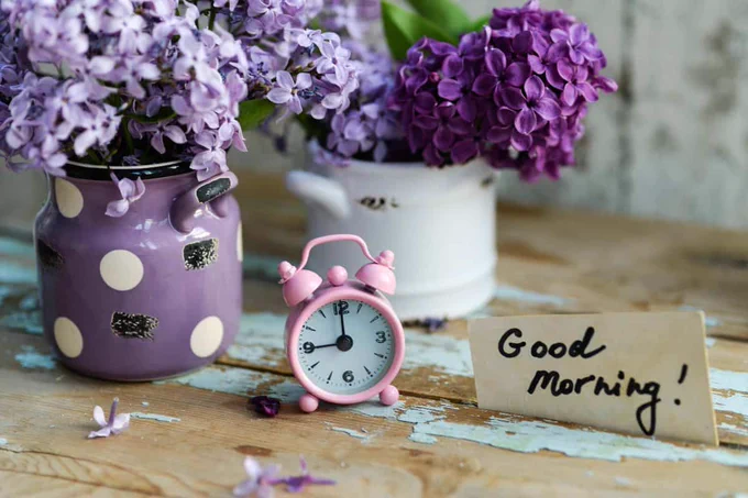 Pink alarm clock on a rustic table beside purple flowers and a handwritten sign that says “Good Morning!”