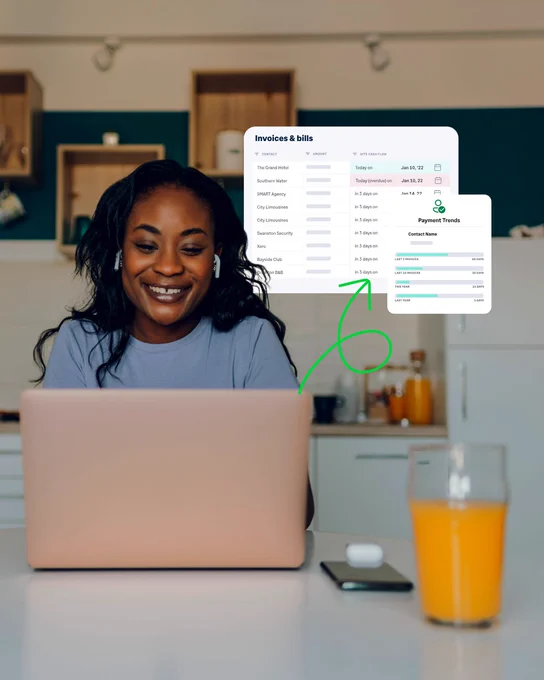 Person seated at a kitchen counter using a laptop. A floating software interface appears above the laptop showing an ‘Invoices & bills’ dashboard with a list of invoices, payment statuses, and a ‘Payment Trends’ chart. A green arrow highlights the dashboard. On the counter are a smartphone, wireless earbuds case, and a glass of orange juice. Modern kitchen cabinets and shelves are visible in the background.
