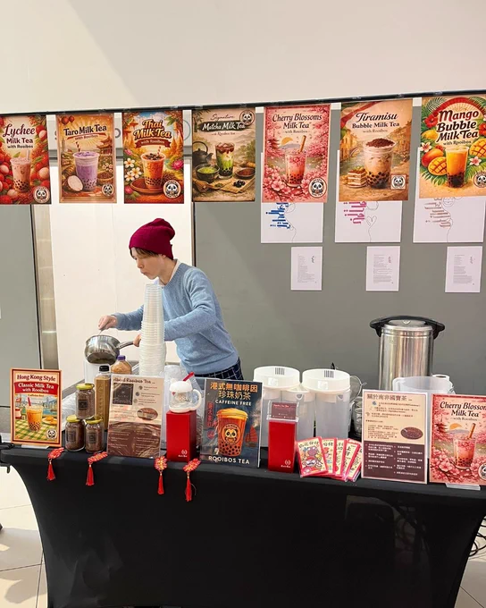 A person wearing a red beanie and blue sweater is preparing drinks at a booth adorned with various tea posters, including flavors like Taro Milk Tea and Mango Bubble Milk Tea. The table is set with drink containers, a menu, and promotional materials from "Little Fluffy Head Cafe.