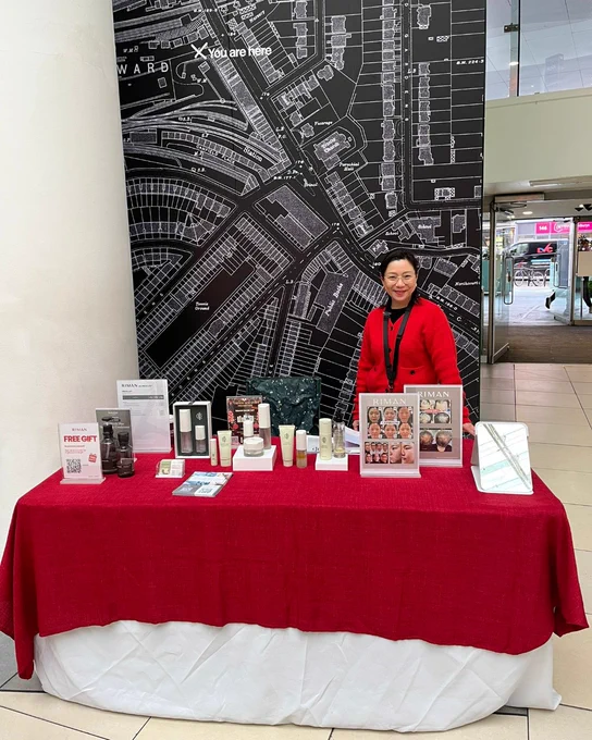 A person stands behind a table covered with a red cloth, displaying various skincare products. The backdrop is a large black and white map labeled "You are Here." The setting appears to be an indoor shopping area.