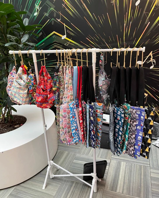 A display rack holds colorful, patterned tote bags with various designs, including some featuring Nintendo characters. The background has a dynamic pattern with radiating lines, and a large green plant is placed next to a curved white bench.