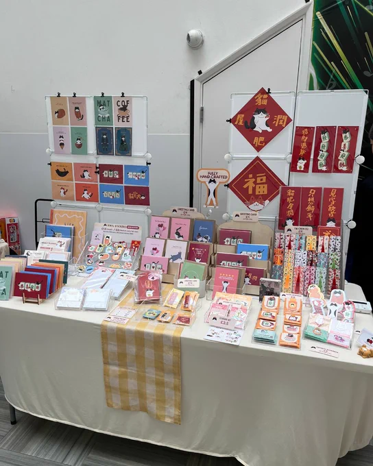 A display at a market stall features colorful notebooks, bookmarks, and decorative wall hangings with illustrations of cats and other designs. There are also Chinese New Year decorations with traditional Chinese characters. The setup is on a table with a white cloth and a yellow checkered runner. The arrangement is neatly organized, showcasing a variety of stationery items.