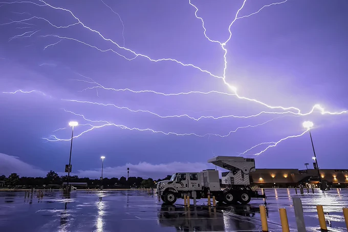 A large lightning storm illuminates the night sky over a parking lot. A utility truck with multiple wheels is parked in the foreground. The area is wet from rain, with glowing streetlights reflecting off the ground.