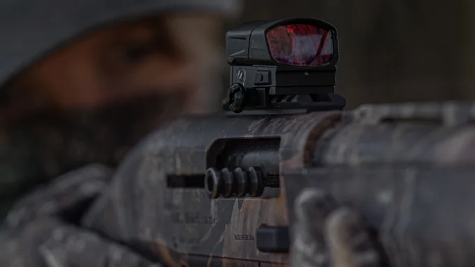 A person is aiming a camouflaged Beretta firearm equipped with a holographic sight. The person's face is partially covered and out of focus in the background.