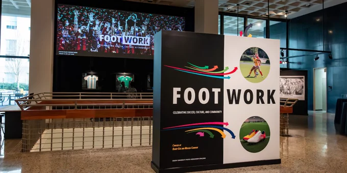 This image shows an exhibition area with a large display sign that says "Footwork: Celebrating Soccer, Culture, and Community." The display features colorful graphics of soccer-related images. In the background, a large screen displays the word "Footwork" over an image of cheering soccer fans. 