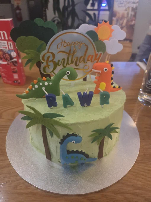 A cake with green icing, decorated with cartoon dinosaurs, trees and clouds