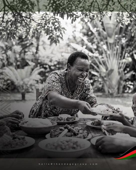 A black and white photograph of an African grandmother mid-laugh, leaning forward across an outdoor wooden table laden with plates of food, passing a full plate to a child whose hands reach toward her from the right of the frame. She wears a patterned dress and a thin necklace. Lush tropical garden foliage fills the background. African geometric chevron patterns mark the left border. A rendition of the Pan-African Flag, with visual influence from the Kenyan flag, in the lower right corner. This image was chosen because it holds the full weight of what African wholesomeness means in practice: abundance given freely, joy shared without ceremony, the ubuntu spirit as daily architecture. The Ndege Foundation© | The Ndege Group® | Africa’s Sovereign Development Trust® (ASDT) | hello@thendegegroup.com