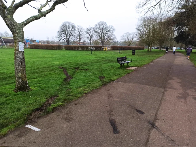 Tyre marks have been left in the grass with mud spread onto the hard surfaced multiuser route through Beddington Park. To the left is a tree yet to produce it's leaves for spring, mid ground is a bench and in the background is an outdoor gym and kids play area.