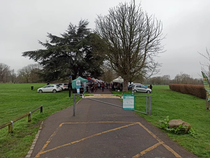 This photo is taken from the east side of the emergency gate near the Pavilion Cafe where a pop up market has been set up with cars parked on the grass and stalls set up either side of the path. People are looking at the wares for sale and also blocking a signposted cycling route. No signage or barriers have been put in place to warn approaching cyclists of the hazard.