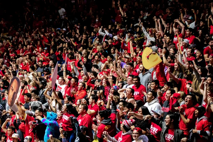 A large crowd of enthusiastic Lobo fans wearing red shirts, many with "UNM" printed on them, are cheering energetically at a basketball game. Some are holding signs and waving foam fingers in celebration. The atmosphere appears lively and packed with excitement.