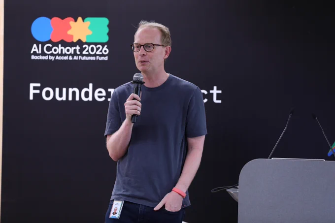 A person speaks into a handheld microphone in front of a black backdrop displaying a colorful logo and the text "AI Cohort 2026 Backed by Accel & AI Futures Fund". A grey podium and two thin microphone stands are partially visible on the right.