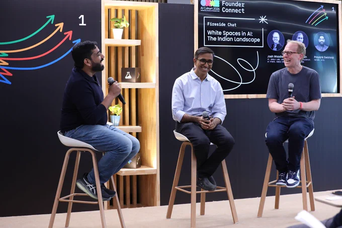 Three people participate in a lively fireside chat on a stage, sitting on high stools and holding microphones while laughing. Behind them, a screen displays text for "Founder Connect AI Cohort 2026" and the session title "Fireside Chat: White Spaces in AI: The Indian Landscape."