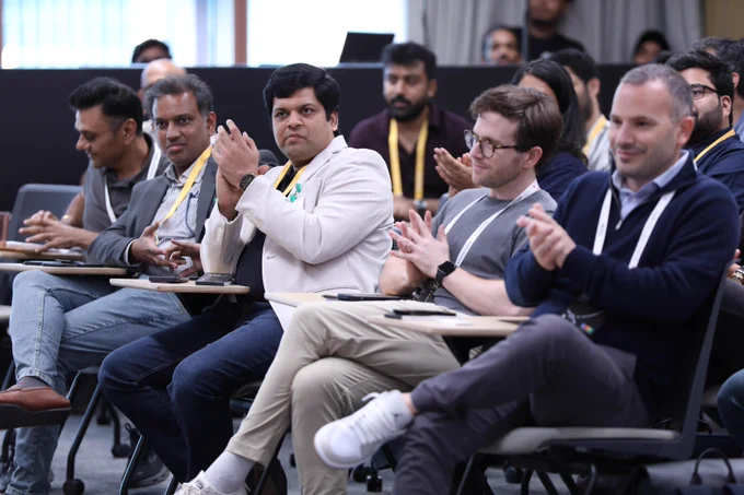 A group of people sit in a row of chairs with attached desks at a professional event, smiling and clapping their hands.