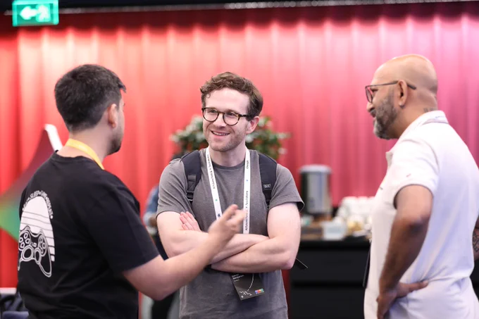 Three people converse at a networking event in front of a red slatted wall.