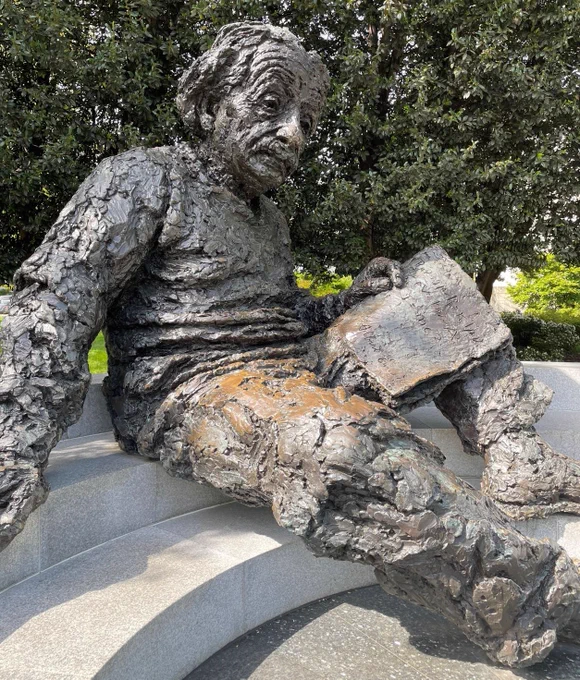 A large metal statue of Albert Einstein holding several papers and sitting on a low curved wall in an outdoor memorial.