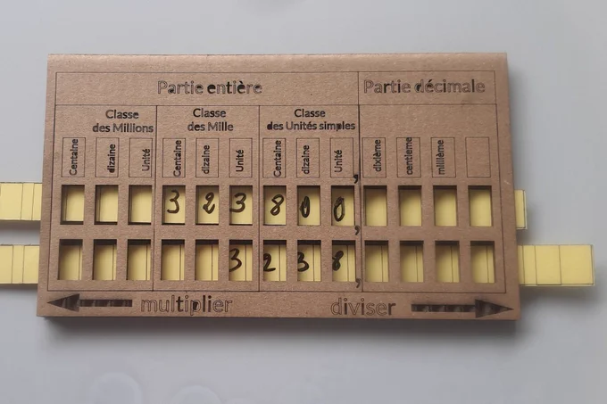 Tableau pédagogique en carton, représentant un tableau de numération. Il est divisé en "partie entière" (classes des millions, des milles, et des unités simples) et "partie décimale" (dixièmes, centièmes, millièmes). Des chiffres sont inscrits dans certaines cases. En bas des flèches indiquent "multiplier" vers la gauche et "diviser" vers la droite
