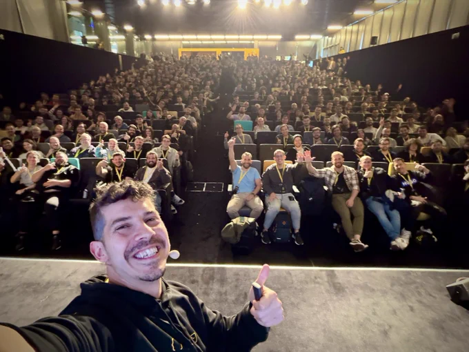 Midudev haciéndose un selfie desde el escenario de la JSConf, sonriendo y levantando el pulgar mientras detrás se ve un auditorio enorme completamente lleno de asistentes mirando a cámara, muchos saludando con la mano. La sala está iluminada con luces cálidas en el techo y el ambiente transmite esa energía de “solo venía a dar una charla” pero parece el final bueno de una película de developers.