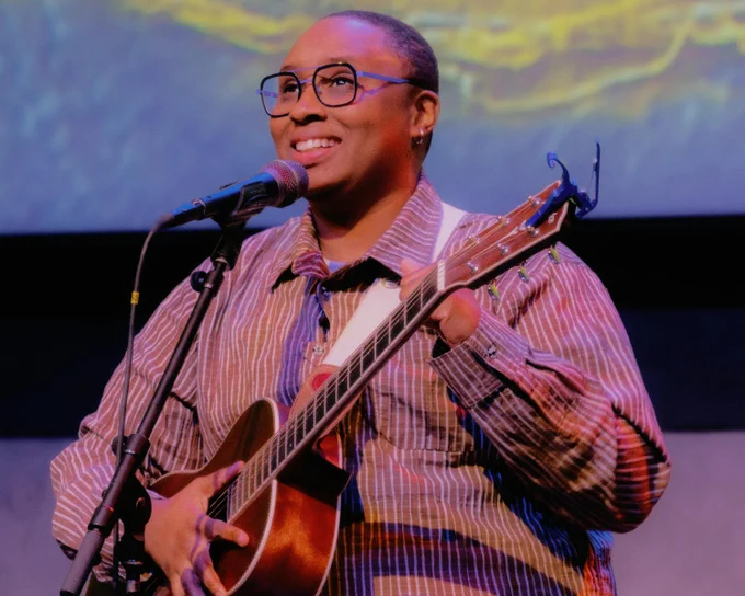 A person stands on stage, smiling while holding an acoustic guitar and singing into a microphone. They are wearing glasses and a striped shirt. The background features colorful, abstract lighting.