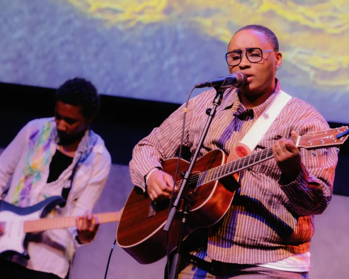 Two musicians performing on stage. The closest individual is playing an acoustic guitar and singing into a microphone. The other musician in the background is playing an electric guitar.