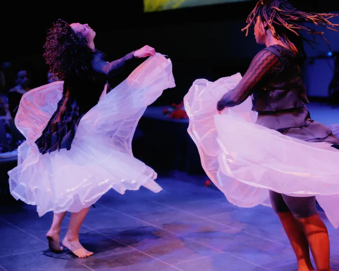 Two performers dance energetically on stage, wearing flowing white skirts that create a dynamic motion effect. The lighting casts a colorful glow, enhancing the sense of movement.
