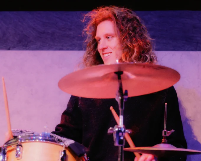 A person with curly hair playing a drum set, smiling, with drumsticks in hand. The background is a simple two-tone wall.