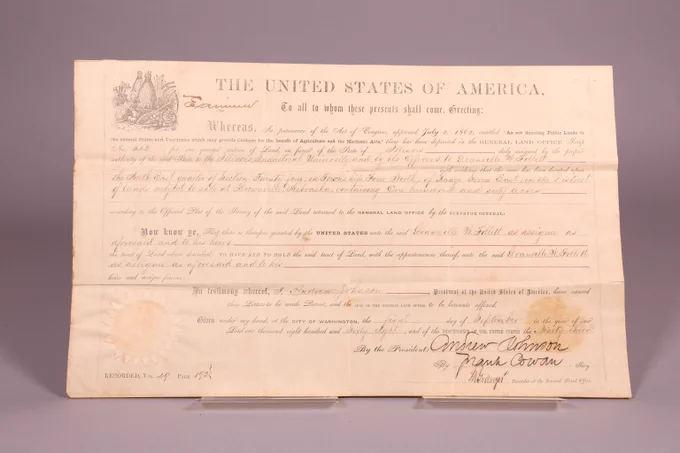 Morrill Act Land patent on stained yellowing paper with printed and handwritten black ink. States tract of land has been issued to Granville M. Follett of Nebraska on September 1st, 1868.  In the bottom left corner is a sun-shaped seal with an eagle in the middle. An ornamental fall harvest illustration of a fall harvest scene adorns the top left corner.