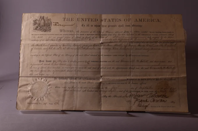 Morrill Act Land patent on stained yellowing paper with printed and handwritten black ink. States tract of land has been issued to Granville M. Follett of Nebraska on September 1st, 1868.  In the bottom left corner is a sun-shaped seal with an eagle in the middle. An ornamental fall harvest illustration of a fall harvest scene adorns the top left corner. Darkened lighting shows prominent horizontal and vertical crease marks and illuminates edges of sun-shaped seal in bottom left corner.