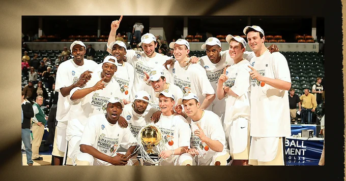 A jubilant basketball team poses on the court with a championship trophy. The players are wearing matching white shirts and caps, indicating their victory. They're celebrating their win, some holding up index fingers. The setting appears to be a professional sports arena.