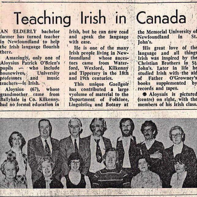 Black and white newspaper clipping with the headline Teaching Irish in Canada. The article text describes an elderly bachelor farmer in Newfoundland who became a teacher of the Irish language. Beneath the text is a group photograph showing several men and women arranged in two rows, some standing and others seated. The people wear formal or semi-formal clothing such as suits, ties, sweaters, and blouses, and face the camera in a posed class portrait. The image appears printed on aged newsprint with visible grain and wear.
