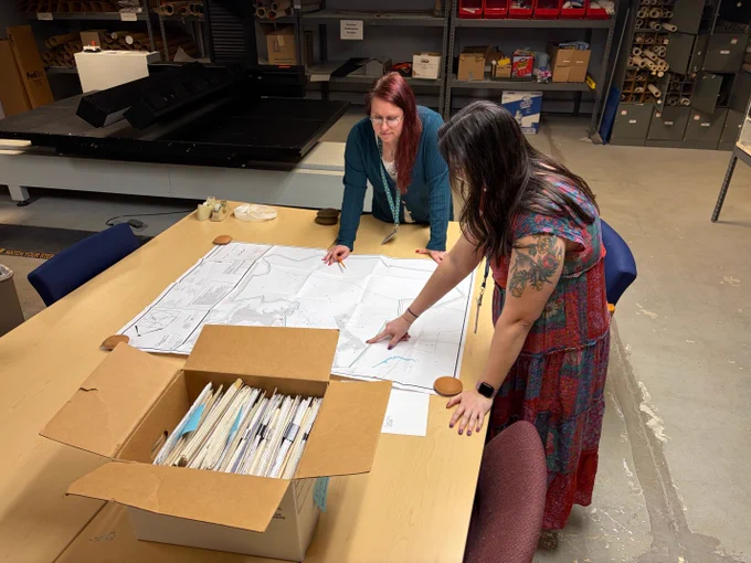Two women examining large architectural blueprints spread out on a table in a storage room filled with shelves and boxes.