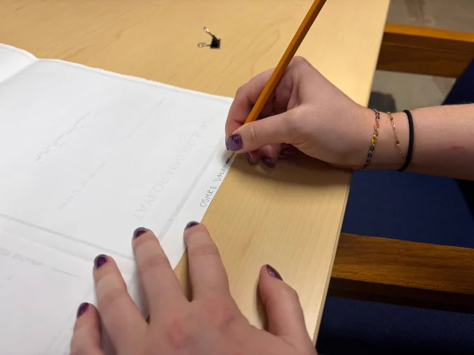 Person writing with a pencil on a white sheet of paper placed on a wooden desk.