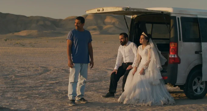 A man and a woman in wedding clothes sit on the back of a truck in the middle of a desert, speaking to a man in casual clothes.