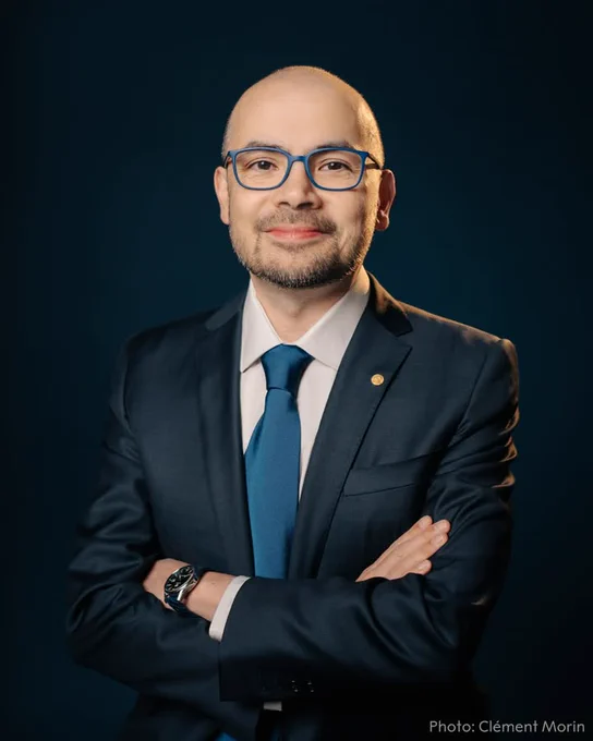 Demis Hassabis wears glasses, a dark suit, and blue tie, standing confidently against a dark blue background.