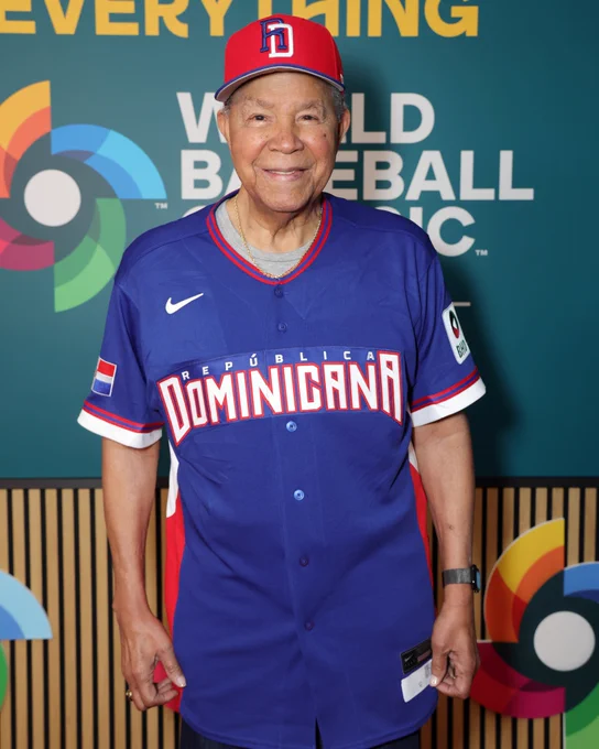 Juan Marichal smiles ahead of the World Baseball Classic semifinal wearing a blue Team Dominican Republic jersey and a red Team Dominican Republic cap.