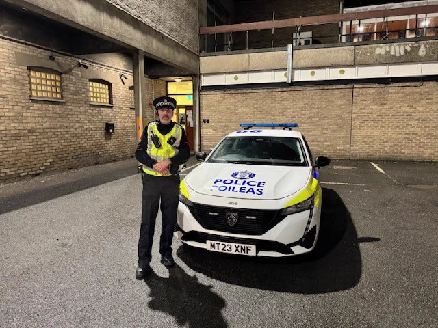 A male police officer in high viz uniform standing in front of a marked police vehicle.