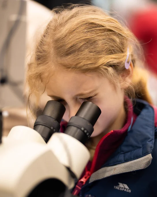 A young girl with light hair is looking through a microscope.