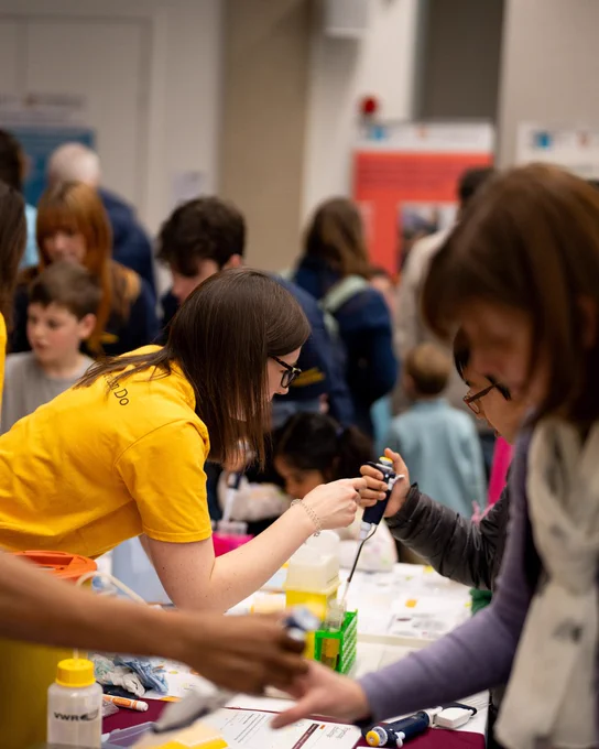 A crowded science fair with attendees of various ages engaging in hands-on activities at a table. A person in a yellow T-shirt assists others with pipettes. In the background, informational posters are visible but not legible.