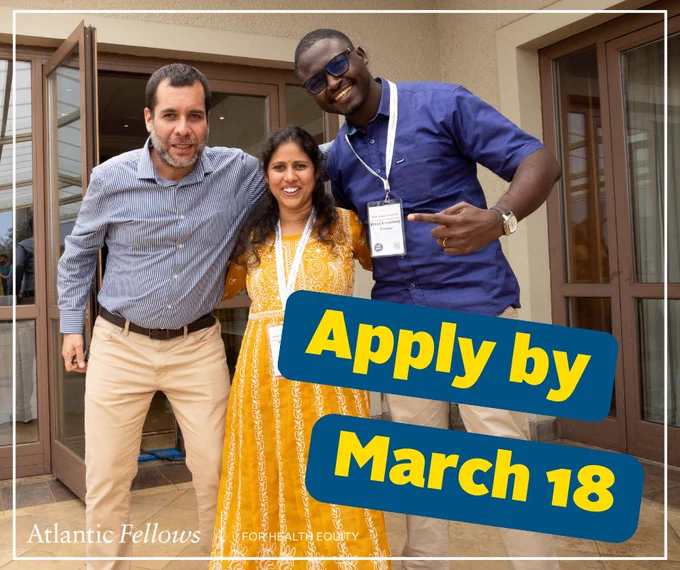 Three people wearing conference badges stand together smiling outside a building with glass doors behind them. Apply by March 18