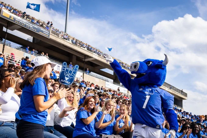 At a stadium, Buffalo's mascot, a blue bull wearing a sports jersey, raises its arm amid cheering fans. Many fans are wearing blue and white clothing. A Buffalo flag is also visible in the background, set against a blue, cloudy sky.
