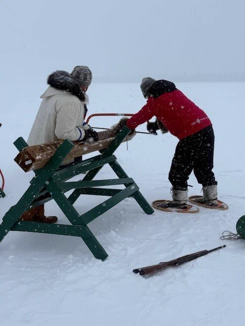 Two people, one in a red parka and one in a white parka are sawing a large log. 