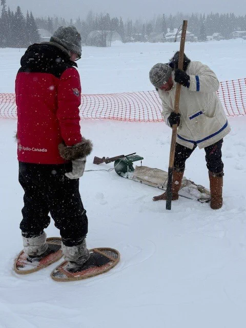 Two people, one in a red parka and one in a white parka, use a large pole to dig into the ice. 