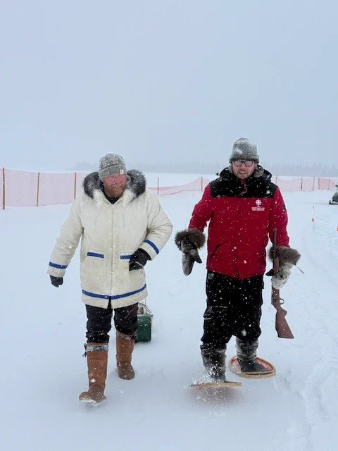 A man in a white parka stands next a man in a red parka wearing snowshoes and holding a rifle.  